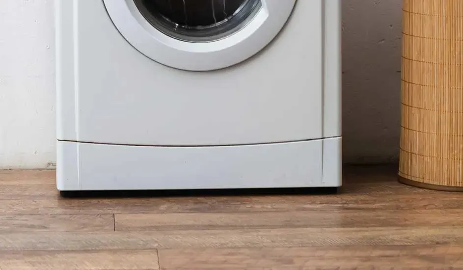 De onderkant van een witte wasmachine op een houten vloer, naast een bamboe wasmand, merk is niet zichtbaar op het apparaat.