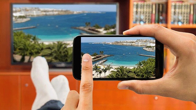 Une main tient un smartphone affichant une plage, avec une télé et une bibliothèque en arrière-plan.