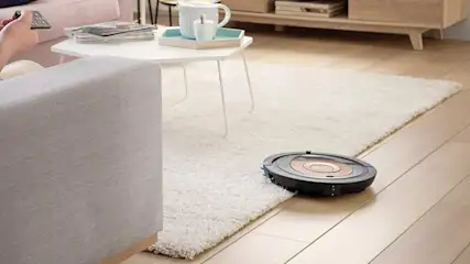 Un aspirateur robot noir nettoie un tapis blanc dans un salon. Une table basse blanche et un canapé sont visibles.
