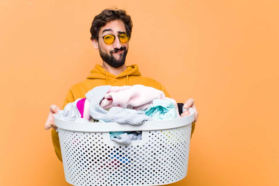 Een witte wasmand vol kleurrijke kleren staat tegen een oranje achtergrond. De mand is van plastic met gaatjes.