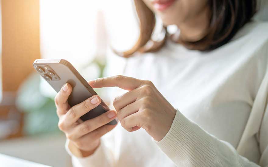 Vrouw in lichte trui gebruikt ontspannen haar smartphone, tikt met één vinger op het scherm terwijl zacht daglicht langs haar gezicht valt.