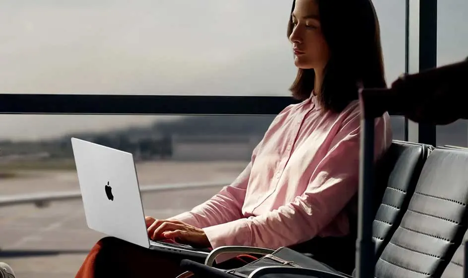 Une femme en chemise rose travaille sur un MacBook, assise à l’aéroport près de grandes fenêtres donnant sur la piste extérieure.