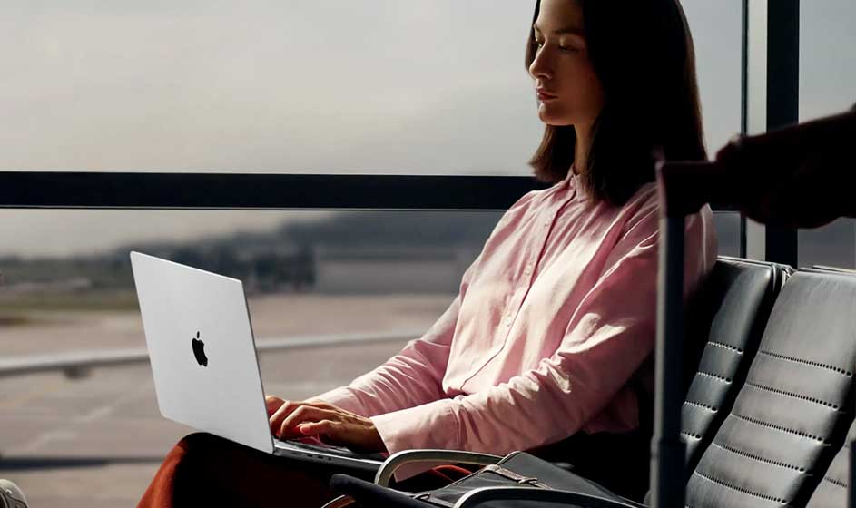 Een vrouw in een roze blouse werkt op een MacBook terwijl zij op luchthavenstoelen bij grote ramen en een landingsbaan zit.
