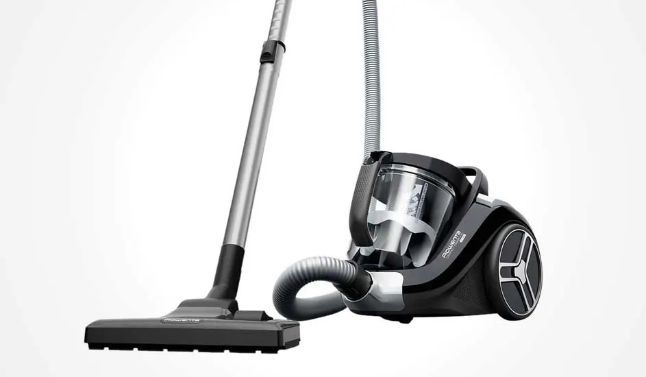 Black cylinder vacuum with clear dust bin, grey hose and metal tube ending in a wide floor head, positioned diagonally on a light background.