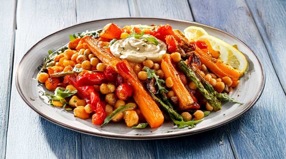 Een bord met kikkererwten, geroosterde wortelen, asperges, paprika, citroen en een witte saus, op een blauwe houten ondergrond.