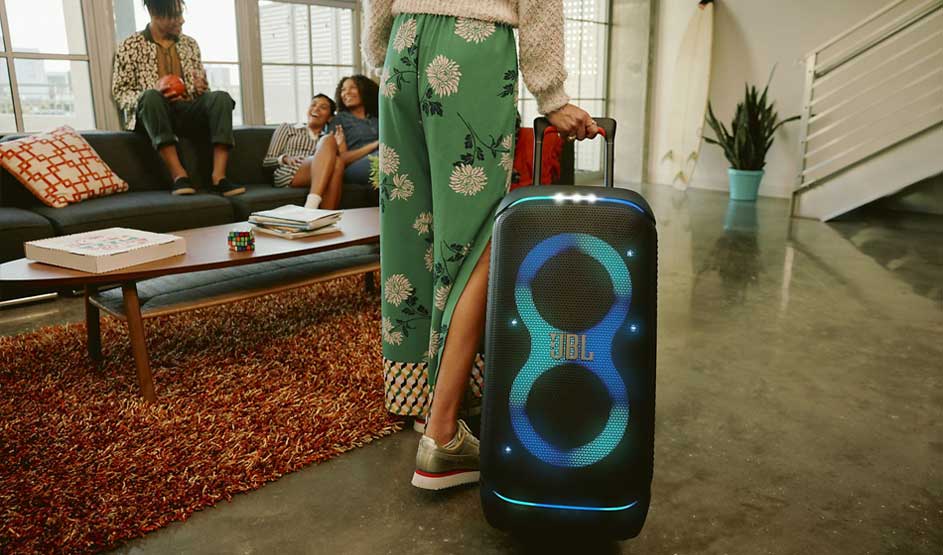 A person pulls a large JBL speaker into the living room while friends sit laughing on the sofa in a cosy setting.