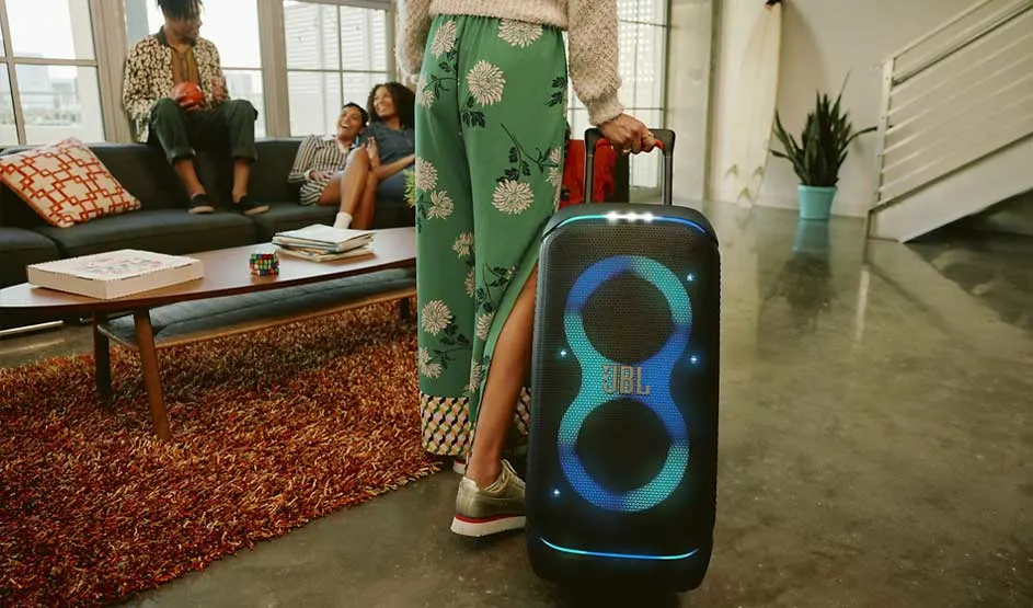 Een persoon trekt een grote JBL-speaker de woonkamer in, terwijl vrienden lachend op de bank zitten in een gezellige setting.