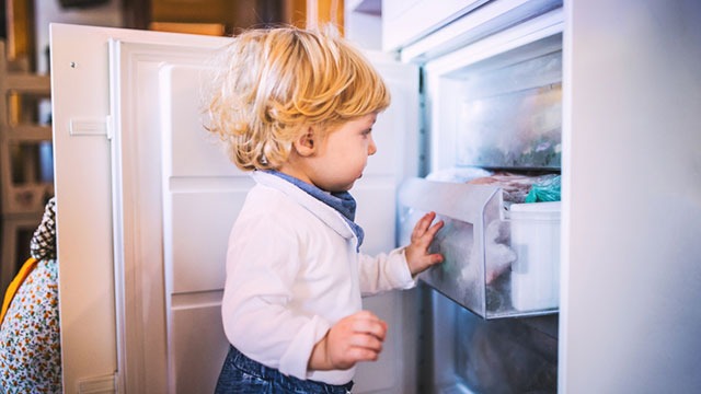 Un enfant ouvre un réfrigérateur blanc. Une étagère est visible avec des articles à l'intérieur. L'intérieur est froid et lumineux.