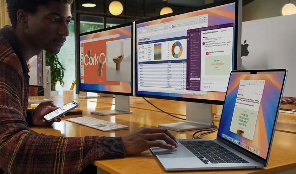 Apple MacBook is connected to two large screens, showing multiple apps; person is also using an iPhone, Apple logo visible.