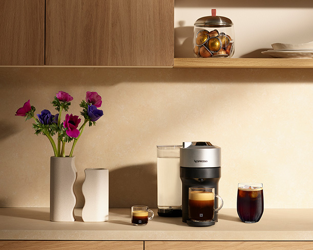 Een keuken met een koffiezetapparaat, vazen met bloemen, glazen met koffie en een pot met koffiecapsules op een plank.