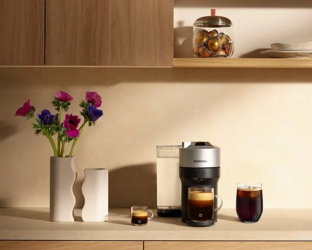 Cuisine avec machine à café, fleurs, verres, et pots sur un comptoir en bois sous des armoires.