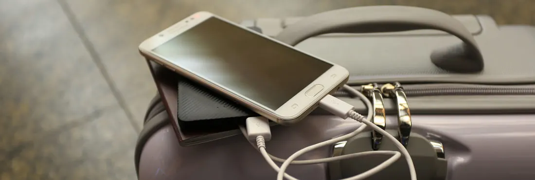 Un téléphone blanc est posé sur une batterie externe noire, tous deux sur une valise grise avec une poignée. Un câble les connecte.