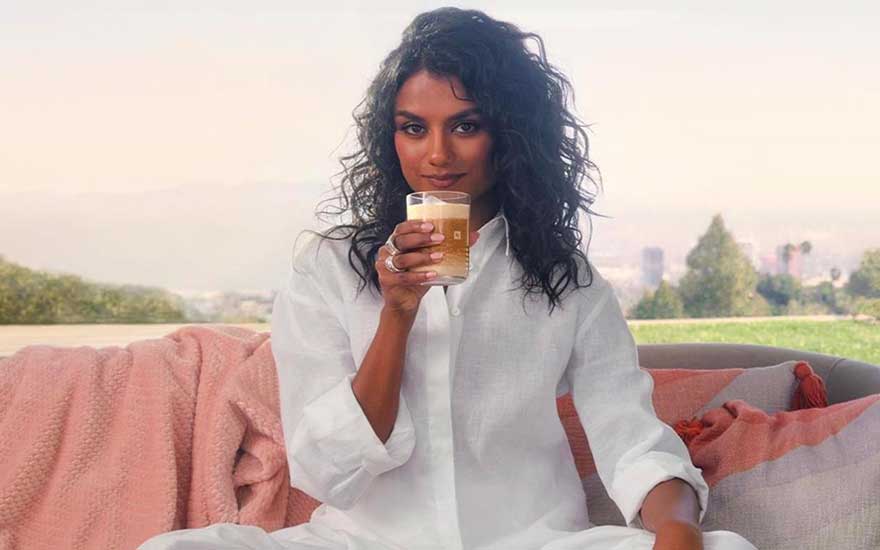 Femme aux cheveux bouclés en chemise blanche savoure son café sur canapé avec plaid rose et vue urbaine.
