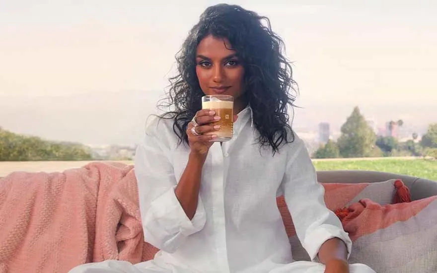 Femme aux cheveux bouclés en chemise blanche savoure son café sur canapé avec plaid rose et vue urbaine.