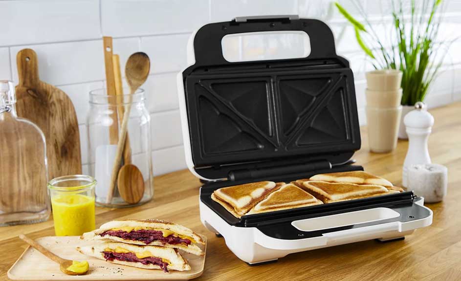 Een wit sandwichapparaat met zwarte binnenkant, met vier driehoekige sandwiches erin. Houten achtergrond met keukengerei.