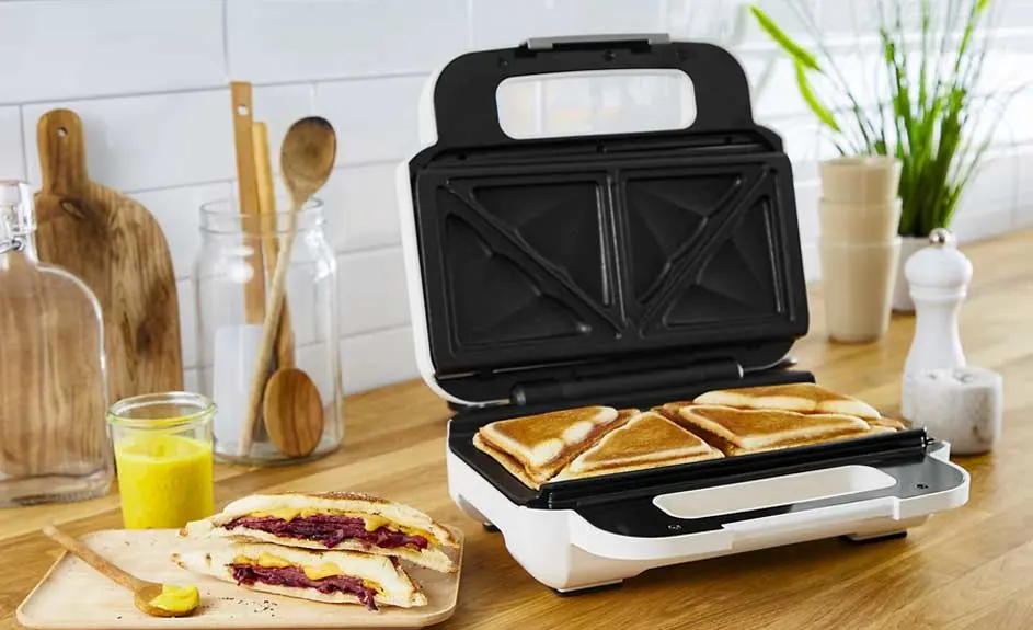 Een wit sandwichapparaat met zwarte binnenkant, met vier driehoekige sandwiches erin. Houten achtergrond met keukengerei.