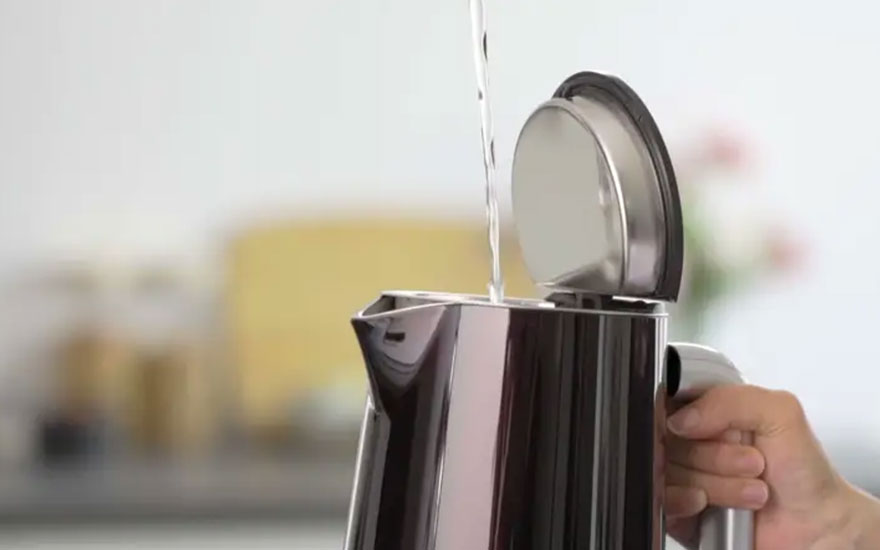 Water wordt in een open zilveren waterkoker gegoten, die op een aanrecht staat.