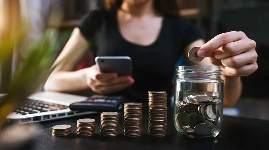 Une personne met des pièces dans un bocal en verre. Des piles de pièces, un ordinateur portable et une calculatrice sont sur une table.