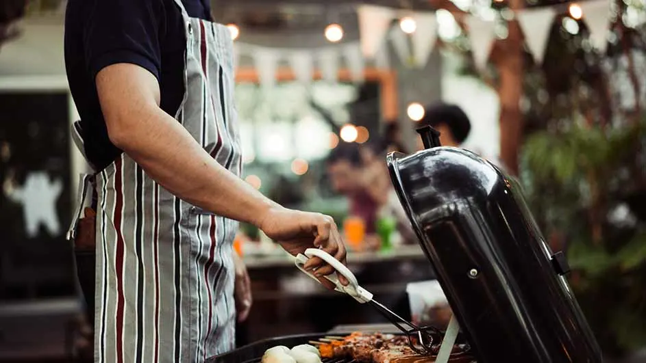 Een persoon met een gestreept schort gebruikt een tang om te barbecueën. Er staat een zwarte barbecue op de voorgrond, met lichten op de achtergrond.
