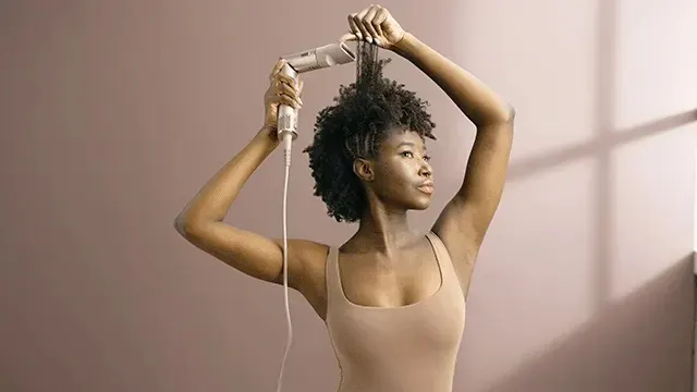 Une personne utilise un sèche-cheveux argenté sur ses cheveux noirs bouclés devant un mur beige avec une lumière naturelle.