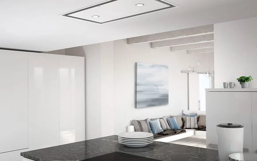 Cuisine moderne blanche avec îlot noir, plaques blanches empilées, hotte intégrée au plafond et coin salon en arrière-plan.