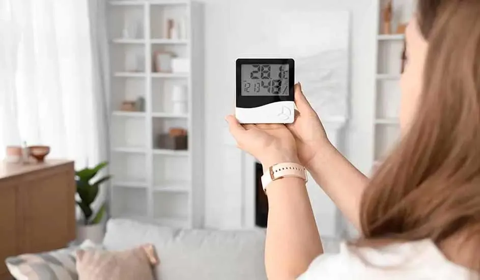 Een rechthoekige, zwart-witte digitale thermometer geeft 28,1 graden aan. Op de achtergrond een woonkamer met witte kasten.