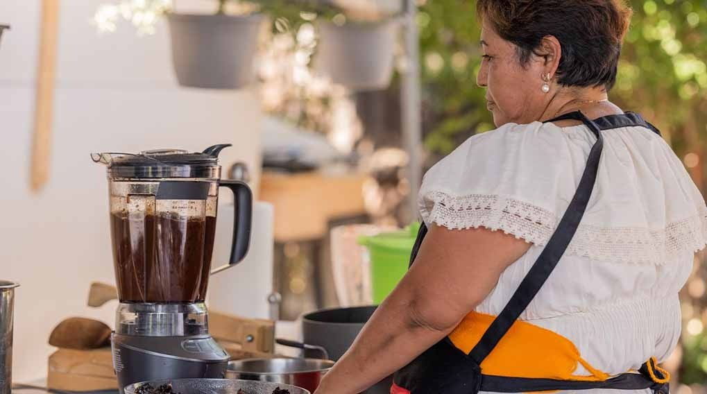 Een blender met een donkere vloeistof staat op een tafel met keukengerei, waaronder pannen en kommen. Achtergrond: groen gebladerte.