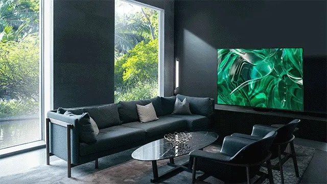 Een woonkamer met een grijze zetel, een glazen tafel, twee stoelen en een grote tv met een groen scherm. Buiten zijn bomen zichtbaar.