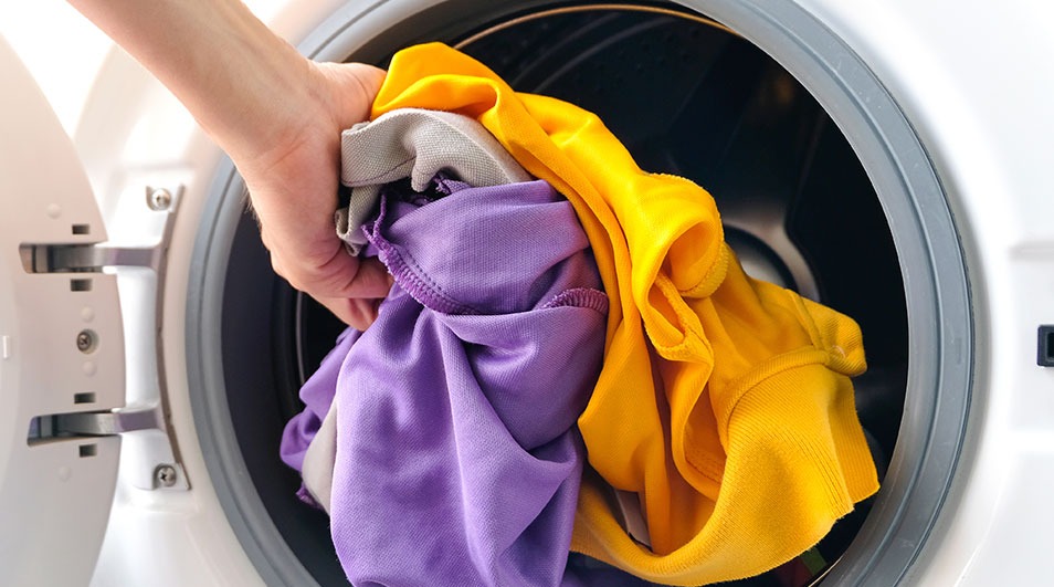 Une main met du linge (jaune, gris, violet) dans un lave-linge blanc avec une porte grise.