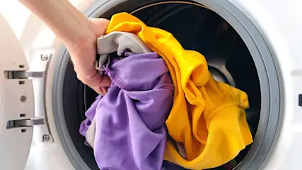 Een hand die kleurrijke kleding in een witte wasmachine plaatst. Gele, paarse en grijze stoffen zijn zichtbaar.
