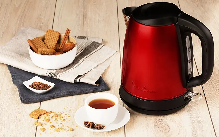 Une bouilloire rouge près d'une tasse de thé, biscuits, et épices sur une table en bois.