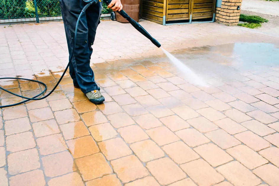 Une personne nettoie une allée pavée orange avec un nettoyeur haute pression noir. L'eau jaillit sur les pavés mouillés.