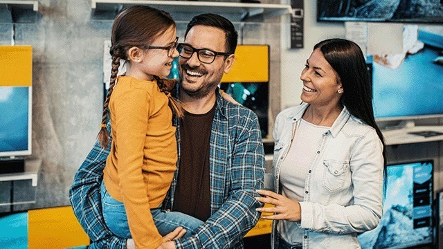 A brightly lit store displays TVs. A man in a blue plaid shirt holds a child in a yellow top. A woman in a white shirt stands nearby.