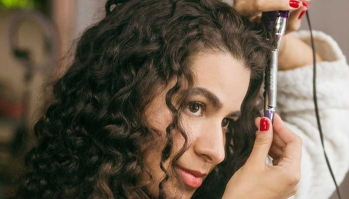 Close-up of a curling iron styling dark curly hair, with red nail polish visible on the hand holding the tool.