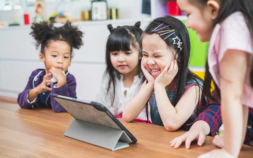 Four children are gathered around a table, watching a tablet placed in a case. They are smiling and seem to be enjoying themselves.