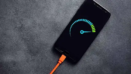Un téléphone noir en charge affiche une jauge bleue et verte sur un fond gris texturé. Un câble orange est connecté au téléphone.