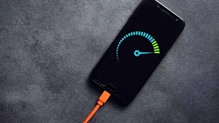 Een zwarte smartphone ligt op een grijze stoffen achtergrond, met een oranje oplaadkabel en een batterij-indicator op het scherm.