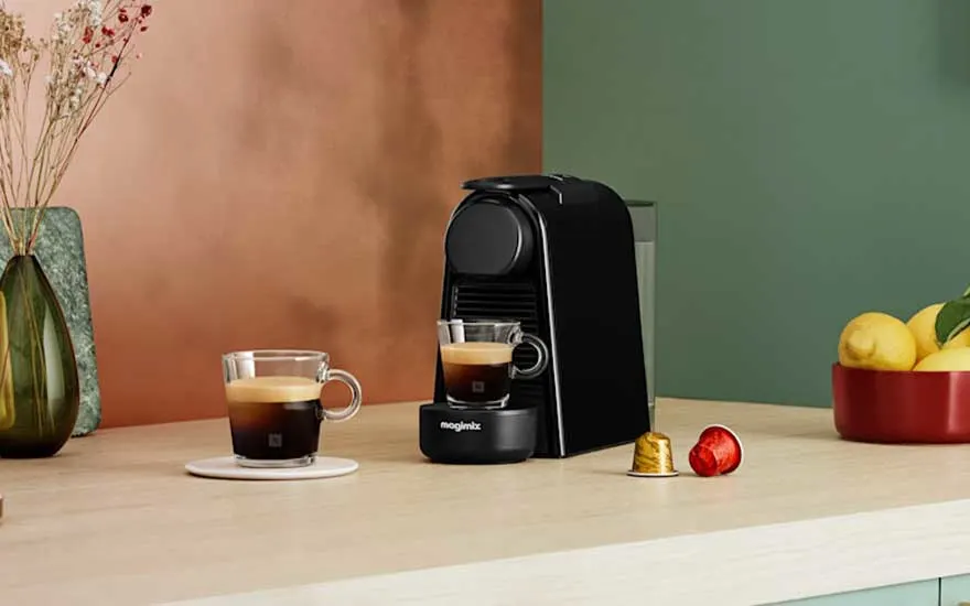Machine à café Magimix noire sur plan de travail avec tasse d'espresso, capsules et fruits dans cuisine chaleureuse.