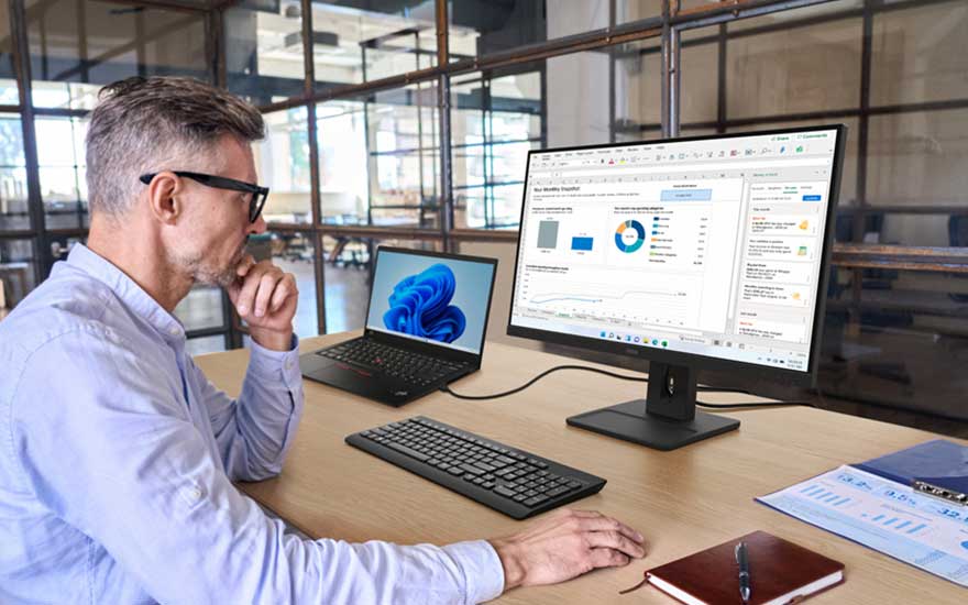 Een man analyseert bedrijfsgegevens op een groot scherm naast een laptop, toetsenbord en notitieboek in een lichte, moderne werkruimte.