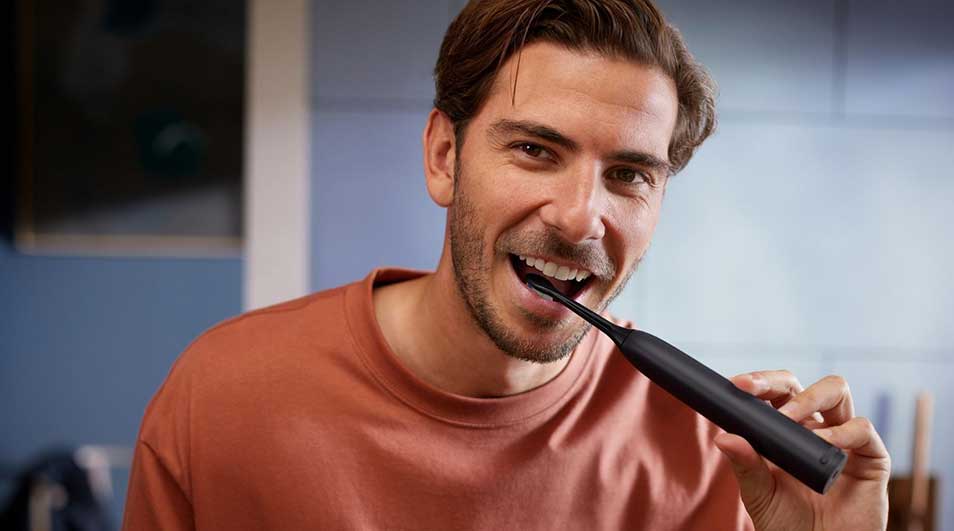 Een man in een rood shirt poetst zijn tanden met een zwarte elektrische tandenborstel in een modern ingerichte badkamer.
