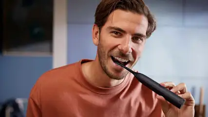 Een man in een rood shirt poetst zijn tanden met een zwarte elektrische tandenborstel in een modern ingerichte badkamer.