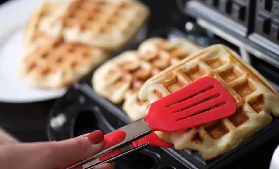 Gaufres dans un gaufrier noir, une spatule rouge les manipule. D'autres gaufres sont sur une assiette blanche à l'arrière-plan.