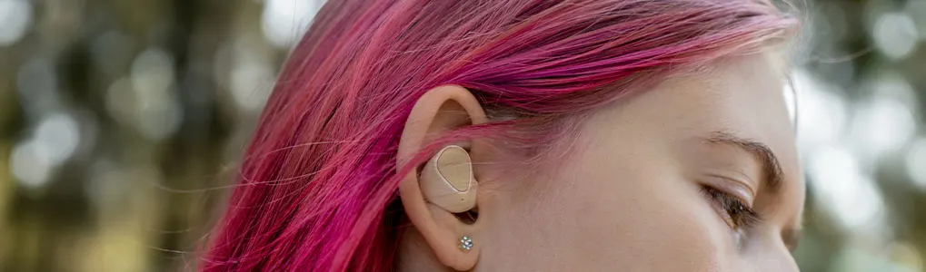 Oreillette beige dans l'oreille, cheveux roses. Fond flou de verdure.