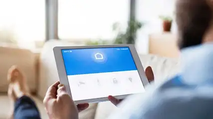 Un écran de tablette affiche une application de maison intelligente bleue et blanche, tenue par des mains devant un canapé beige.