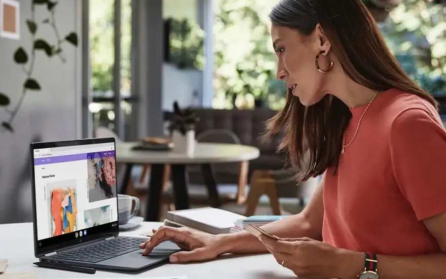 Een laptop op een wit bureau met een kopje, boeken en een smartphone. Een open raam laat daglicht binnen.
