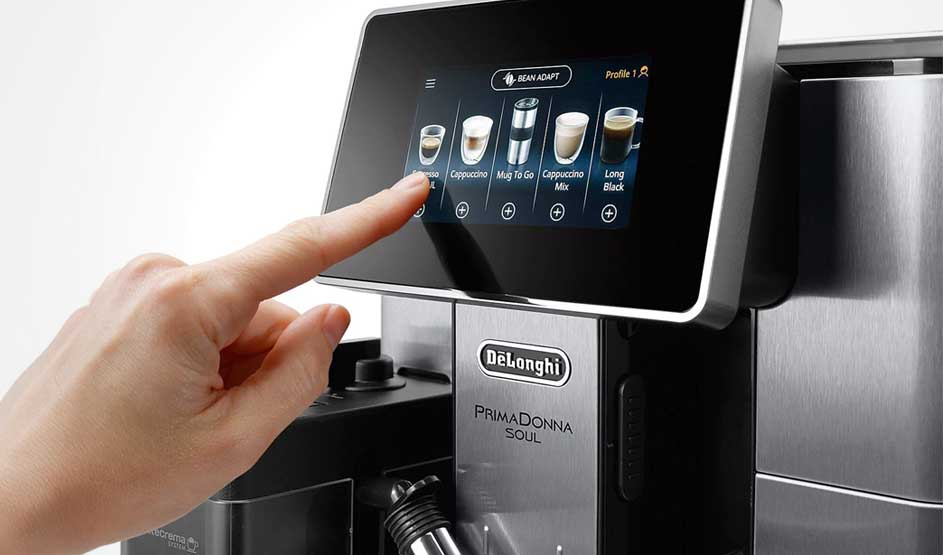A hand taps the large colour touchscreen of a De'Longhi coffee machine, selecting between cappuccino, espresso and several other drinks.