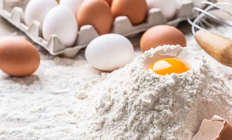 Préparation de pâte : œufs blancs et bruns, farine avec un jaune d'œuf au centre et un fouet en arrière-plan.