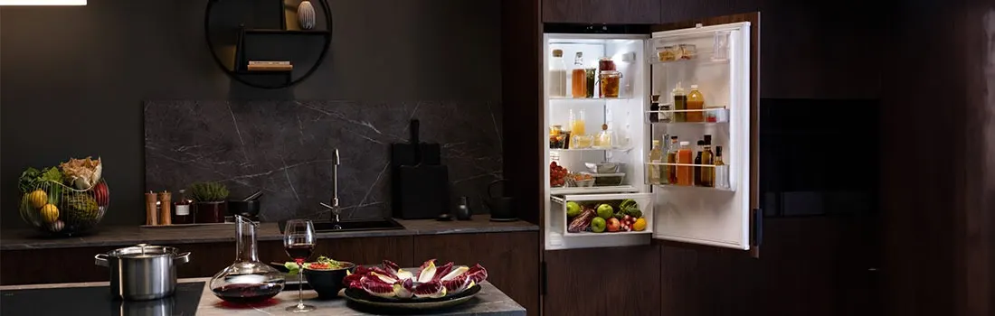 Cuisine sombre avec un réfrigérateur ouvert rempli de nourriture, un évier, des ustensiles de cuisine et des fruits.