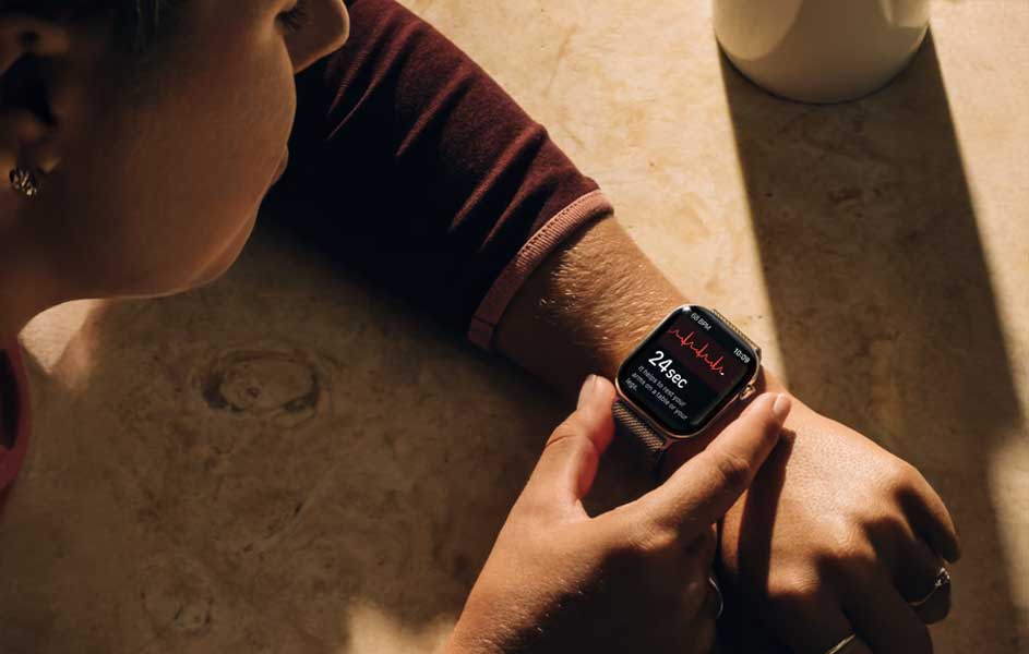 Une personne consulte une montre connectée à son poignet. L’écran affiche des données de santé, tandis que ses bras reposent sur une table en bois près d’une tasse claire.