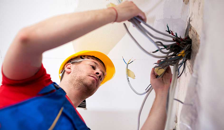 Un électricien en casque jaune manipule des câbles gris sortant d'un mur blanc. L'arrière-plan est un plafond blanc.